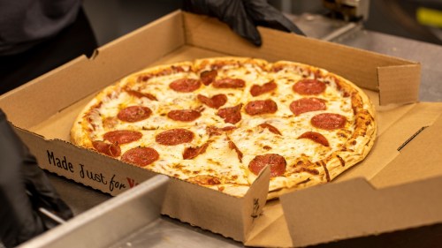 A person in black gloves holds a pepperoni pizza in a cardboard box, labeled "Made Just for You."