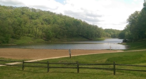 A serene lake surrounded by lush green hills under a partly cloudy sky, with a sandy area in the foreground.