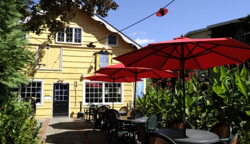A charming yellow house with red umbrellas over outdoor tables, surrounded by greenery and blue sky.