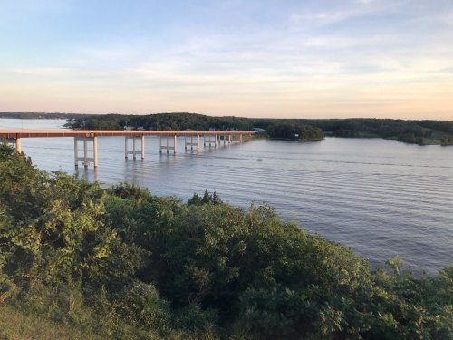 A serene view of a river with a bridge, surrounded by lush greenery and a calm sky at sunset.