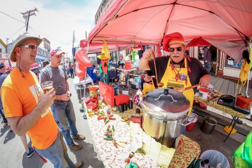 A vendor in a red hat serves food at a lively street festival, with colorful decorations and attendees enjoying the event.