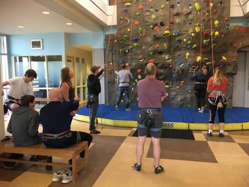 A group of people climbing a rock wall indoors, with some waiting on a bench and others preparing to climb.