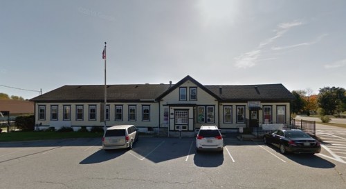 A one-story building with a flagpole, featuring a white and black exterior, surrounded by parked cars.