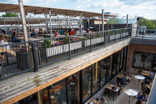 A rooftop dining area with people enjoying meals, surrounded by greenery and modern architecture.