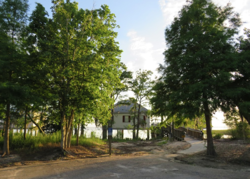 A serene pathway leads to a house by the water, framed by lush green trees under a clear sky.