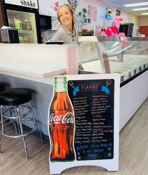 A smiling woman stands behind an ice cream counter with a colorful menu board featuring various flavors.