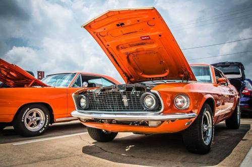 A classic orange muscle car with the hood open, showcasing its engine, parked alongside another orange vehicle.