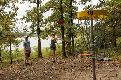 Two people playing disc golf in a wooded area, with a basket in the foreground and a lake visible in the background.