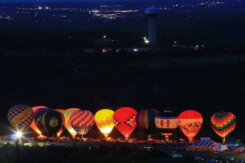 A night scene of colorful hot air balloons glowing against a dark sky, with a distant water tower and city lights.