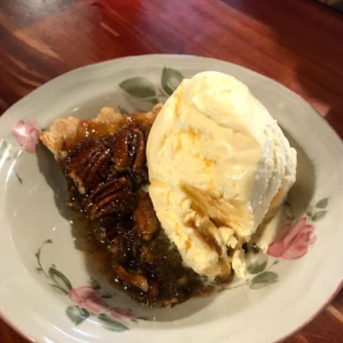 A slice of pecan pie with a scoop of vanilla ice cream on a floral plate, resting on a wooden table.