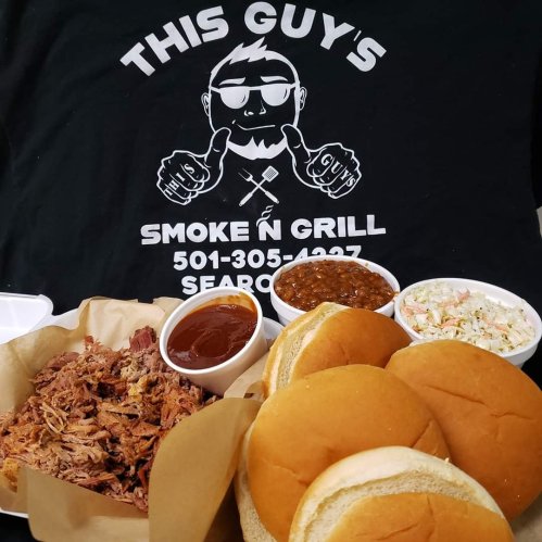 A plate of pulled pork with barbecue sauce, two buns, baked beans, and coleslaw, alongside a black t-shirt advertising a BBQ restaurant.