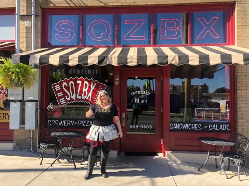 A woman in a black shirt and skirt stands outside SQZBX, a brewery and pizzeria, giving a thumbs up.