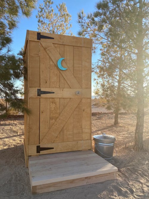 A wooden outdoor toilet with a crescent moon symbol, surrounded by trees and a metal trash can nearby.