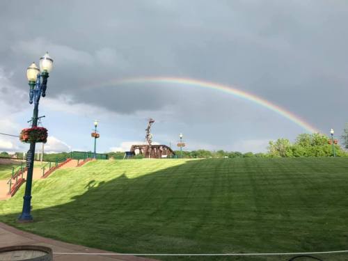 A vibrant rainbow arcs over a green lawn, with a cloudy sky and decorative lampposts in the foreground.