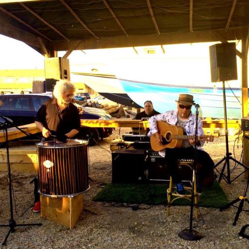 Two musicians perform outdoors under a shelter; one plays guitar while the other uses a drum. A boat is in the background.