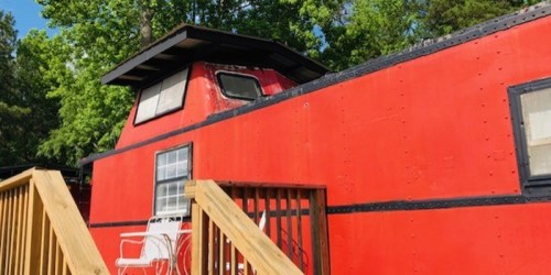 A bright red train caboose converted into a cozy cabin, surrounded by green trees and a wooden deck.