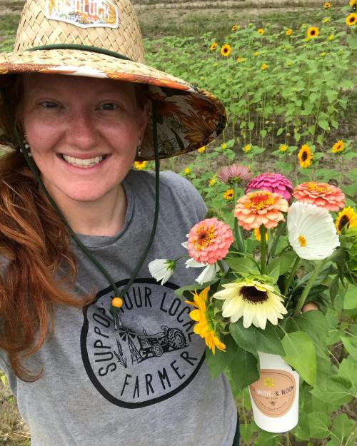 A smiling person in a straw hat holds a bouquet of colorful flowers in a field with sunflowers in the background.