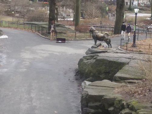 A park scene featuring a dog statue on a rock, with a musician playing nearby and people walking in the background.