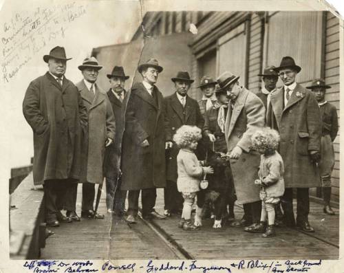 A historic black-and-white photo of a group of men in hats, with two children and a dog, standing on a dock.