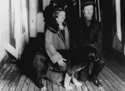 A man and woman sit on a wooden deck with a large dog, both smiling at the camera in a vintage black-and-white photo.