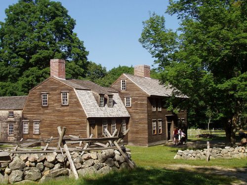 A historic wooden house with a stone fence, surrounded by trees and a clear blue sky. People are visible near the entrance.