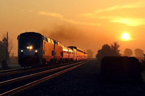 A train travels along tracks at sunset, with a warm orange glow and silhouettes of trees in the background.
