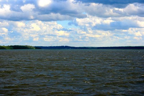 A wide view of a lake with rippling water under a cloudy sky and distant green hills on the horizon.