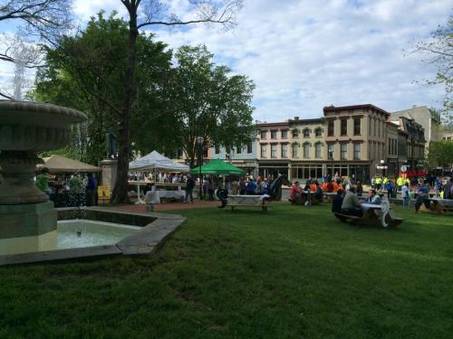 A lively park scene with people gathered around a fountain, tents, and green grass under a partly cloudy sky.