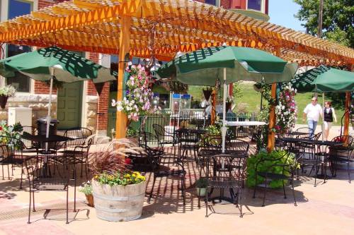 Outdoor dining area with green umbrellas, hanging flowers, and empty tables under a wooden pergola.