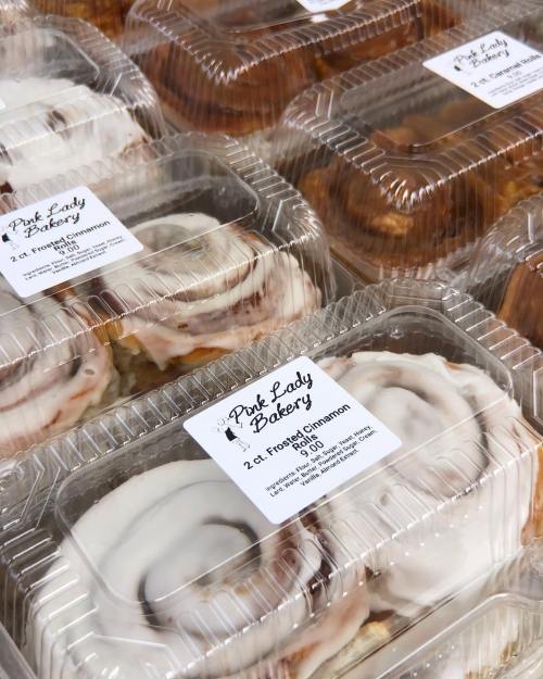 Clear plastic containers filled with frosted cinnamon rolls from Pink Lady Bakery, labeled with pricing and bakery name.