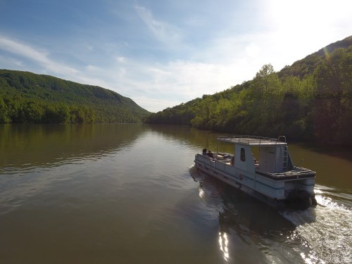 A boat glides along a calm river, surrounded by lush green hills under a bright sky.