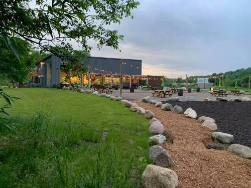 A modern building with outdoor seating, surrounded by greenery and a stone pathway, under a cloudy sky.