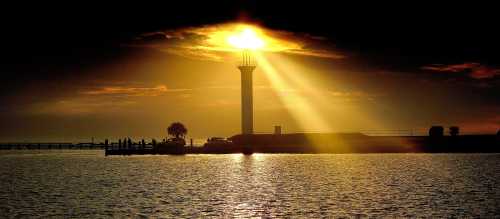 A lighthouse silhouetted against a dramatic sunset, with rays of light shining over calm waters.