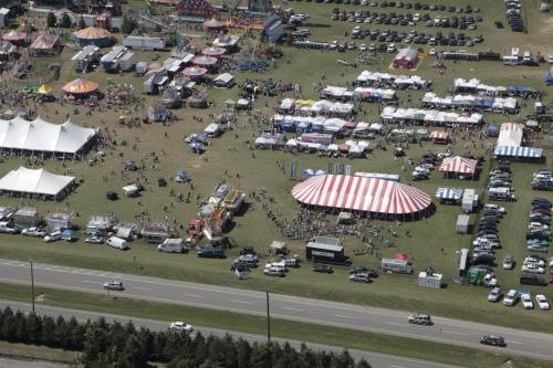Aerial view of a large outdoor fair with tents, rides, and crowds, surrounded by parked cars and open fields.