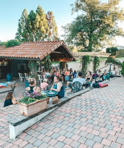 A group of people gathered outdoors at a venue with a tiled roof, surrounded by greenery and flowers, enjoying a social event.