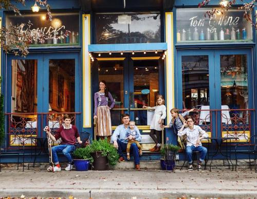 A family poses on the steps of a colorful storefront, smiling and dressed in casual attire, with plants around them.