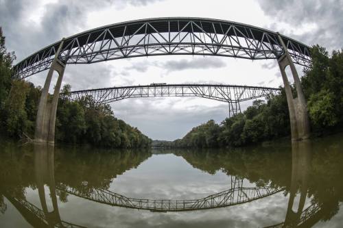 A wide river reflects two large, steel bridges above, surrounded by lush greenery under a cloudy sky.