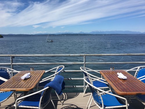 A scenic view of a calm lake with mountains in the background, featuring outdoor dining tables and chairs.