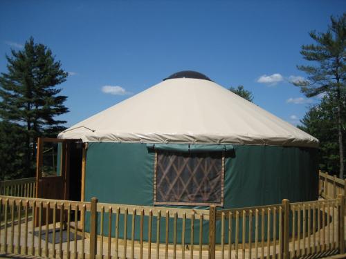 A green yurt with a white dome roof, surrounded by a wooden deck and trees under a blue sky.
