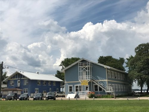 Two blue buildings with a grassy area in front, under a cloudy sky, with parked cars nearby.