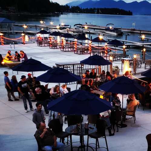 A lively outdoor bar scene by the water, with people socializing under blue umbrellas and string lights at dusk.