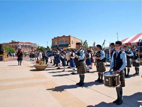 A group of musicians in kilts playing bagpipes and drums at an outdoor event under a clear blue sky.
