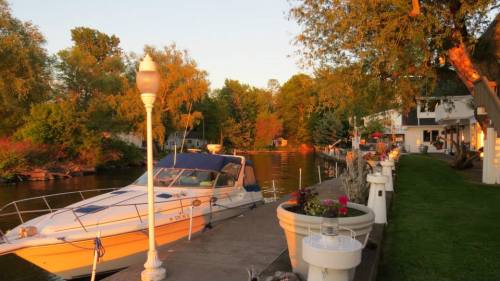 A serene riverside scene at sunset, featuring a boat docked along a walkway lined with flowers and lampposts.