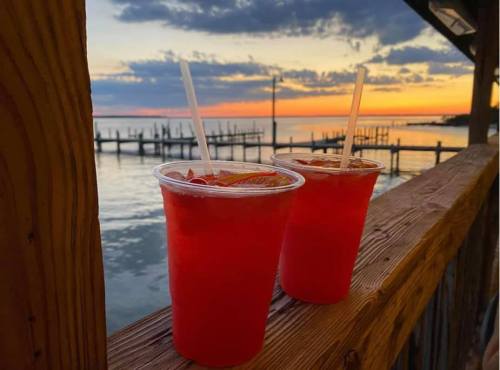 Two red drinks with straws sit on a wooden railing, overlooking a sunset by the water with docks in the background.