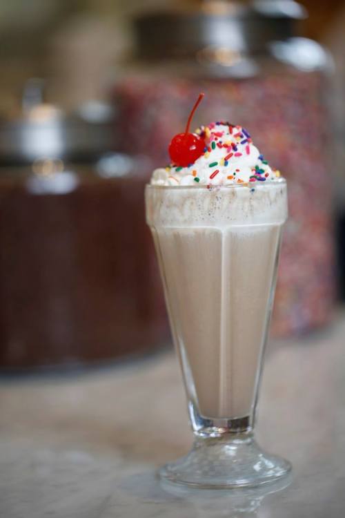 A tall glass of milkshake topped with whipped cream, a cherry, and colorful sprinkles, with jars of toppings in the background.