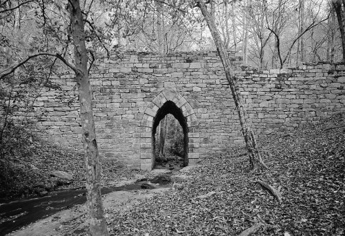 A stone archway bridge surrounded by trees and fallen leaves, with a small stream flowing beneath it.