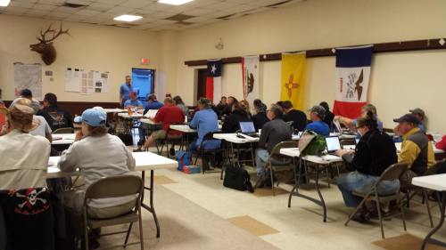 A classroom setting with people seated at tables, engaged in a session, with flags on the walls.