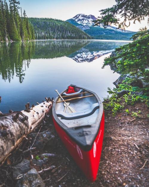 A red canoe rests on the shore of a calm lake, surrounded by trees and mountains reflecting in the water.