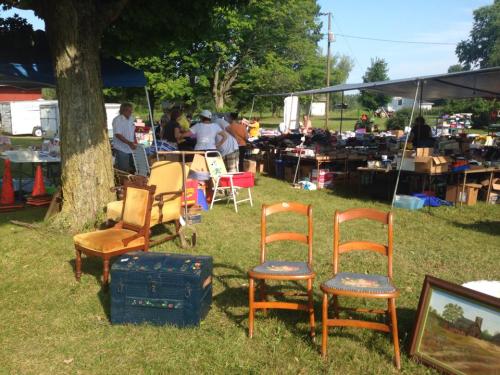 A bustling outdoor flea market with various items for sale, including furniture and boxes, under a blue sky.