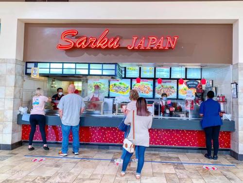 A food counter at Sarku Japan with customers ordering, featuring colorful menu displays and a bright neon sign.